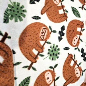 May include: White fabric with a repeating pattern of brown sloths hanging from branches, interspersed with green leaves and floral shapes. The sloths have smiling faces and are holding onto the branches. The fabric has a soft, plush texture.