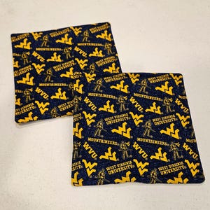 May include: Two square fabric coasters with a navy blue background and a repeating pattern of yellow WVU logos, Mountaineer figures, and text that reads "West Virginia University".