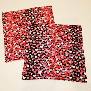 May include: Two square fabric items with a black background covered in a pattern of red, pink, and white hearts. The edges are finished with a red border. The items appear to be quilted or padded.