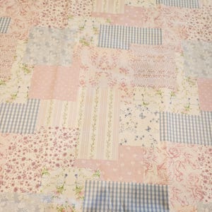 Op de afbeelding: Een patchworkstof met diverse patronen, waaronder lichtblauw-witte vichyruitjes, roze bloemenprints, verticale strepen met kleine bloemetjes en effen lichtroze vierkanten, gerangschikt in rechthoekige en vierkante secties.