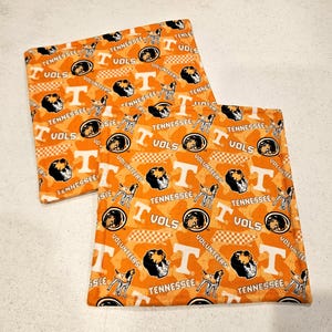 May include: Two square fabric coasters featuring an orange background with a repeating pattern of the Tennessee Volunteers logo, the letter T, and a dog mascot. The design includes the text Tennessee, Vols, and Volunteers in white and black.