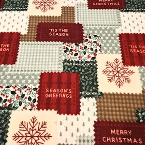 May include: A fleece blanket featuring a patchwork design of red, green, brown, and cream squares. The pattern includes red snowflakes, holly berries, and text reading Season's Greetings, Merry Christmas, and 'Tis the Season on various blocks.