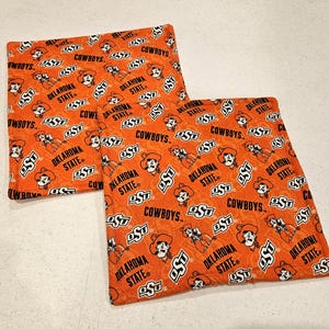 May include: Two square fabric coasters featuring an orange background with a repeating pattern of the Oklahoma State University logo, the text Cowboys, and the mascot Pistol Pete. The fabric has a quilted texture with visible stitching throughout.