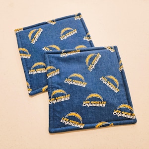 May include: Two square blue fabric coasters. Each coaster features a repeating pattern of the Los Angeles Chargers logo in yellow and white. The coasters are edged with a yellow border.