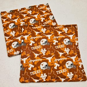 May include: Two square fabric coasters with a repeating pattern of white football helmets, the word "TEXAS", the outline of the state of Texas, and the word "Longhorns" on a brown and orange background.