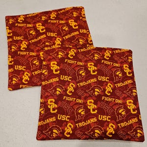 May include: Two square fabric items with a maroon background and a repeating pattern of yellow USC logos, Trojan helmets, and the words "FIGHT ON!" and "TROJANS".
