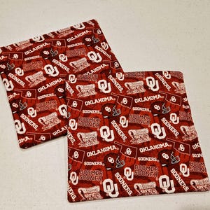 May include: Two square fabric coasters with a repeating pattern of the words "OKLAHOMA SOONERS" and "BOOMER SOONER" in white on a crimson background. The design includes the OU logo and football helmets.