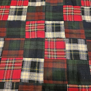 May include: A patchwork quilt constructed from square fabric pieces featuring various plaid patterns in shades of red, dark green, navy blue, and cream. The textile surface displays a repeating grid arrangement of these distinct tartan designs.