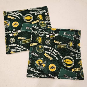 May include: Two square fabric coasters featuring a dark green background with a repeating pattern of North Dakota State University logos, NDSU text, football graphics, bison icons, and the phrases Bison Pride and North Dakota State University.
