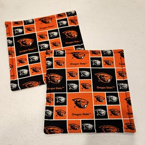 May include: Two square fabric hot pads with a repeating pattern of orange, black, and white squares. Each square features the Oregon State logo and the words "Oregon State". The hot pads are made of a thick, quilted material.