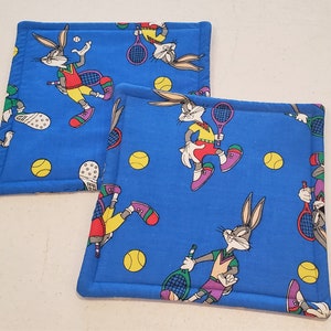 Könnte beinhalten: Zwei blaue Stoffuntersetzer mit weißem Rand zeigen ein Cartoon-Kaninchen, das Tennis spielt. Das Kaninchen trägt ein rot-gelb-blaues Outfit. Die Untersetzer haben einen weißen Rand und sind mit gelben Tennisbällen verziert.