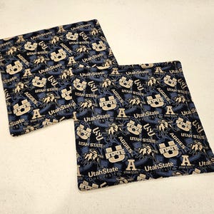 May include: Two square fabric items with a navy blue background and a repeating pattern. The pattern includes the words "Utah State", "Aggies", and the letter "A" in white and tan. The items are likely coasters or small decorative squares.