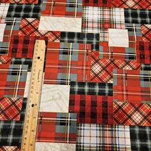 May include: A patchwork fabric with a variety of plaid patterns in red, black, gray, and white. A wooden ruler is placed diagonally across the fabric, showing inch measurements. The fabric design features a mix of textures and colors.
