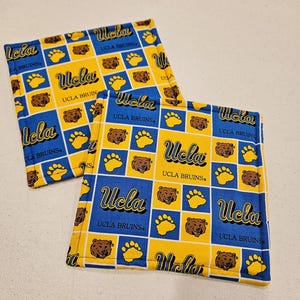 May include: Two square fabric items with a repeating pattern of blue and yellow squares. Each square features a bear paw print, a bear face, and the text "Ucla UCLA Bruins". The items are likely coasters or trivets.