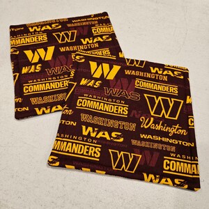 Puede incluir: Dos posavasos de tela marrón con un patrón repetido del logotipo de los Washington Commanders y las palabras "Washington Commanders" y "WAS" en amarillo.