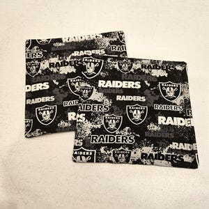 May include: Two black and white fabric pot holders with a repeated pattern of the Las Vegas Raiders logo and the word "RAIDERS".