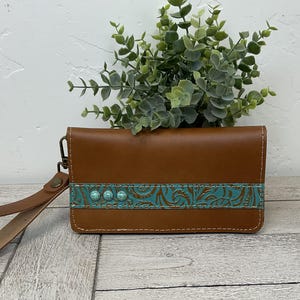May include: A brown leather clutch with a turquoise floral embossed design. The clutch has a wrist strap and three turquoise studs. The stitching is white. The background includes greenery and a light-colored surface.