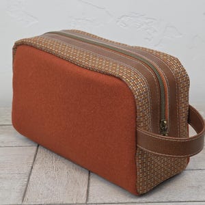 May include: A rectangular toiletry bag with a rust-colored felt body and a woven brown top. The bag features a brown leather handle and zipper pulls. The zipper is a dark bronze color.
