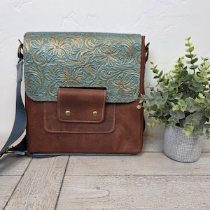 May include: A brown leather crossbody bag with a turquoise floral embossed flap and a small front pocket. The bag has a blue adjustable strap and gold-colored hardware. A small potted plant sits to the right of the bag.