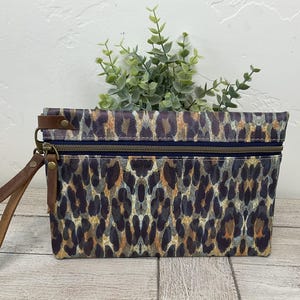 May include: A rectangular clutch bag with a leopard print pattern in shades of brown, tan, and navy blue. The bag has a navy blue zip and brown leather wrist strap. A small green plant sits on top of the bag.