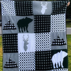 Può includere: Una coperta all'uncinetto in bianco e nero con un motivo a scacchi e vari disegni di animali del bosco e montagne. La coperta presenta un cervo, un orso e cime di montagne.