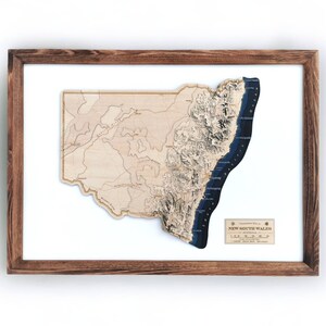 May include: A wooden 3D map of New South Wales, Australia, with a blue ocean and brown mountains. The map is framed in a dark brown wood frame.