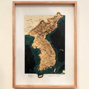 May include: A framed 3D topographic map of the Korean Peninsula featuring layered wood construction. The landmass is rendered in light tan, while the surrounding deep ocean areas are depicted in dark teal and black. Text includes Korea.