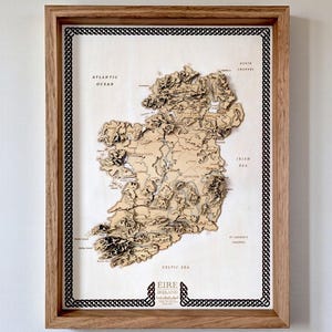 May include: A framed 3D wooden topographic map of Ireland, labelled Eire Ireland, featuring layered terrain details, geographical names, and a black Celtic knot border. The map is encased in a light-coloured wooden frame with a clean, neutral background.