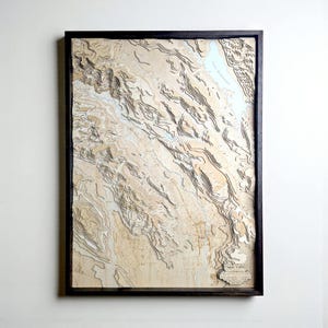 May include: A framed, three-dimensional topographical map of Napa Valley, California. The map is made of layered wood, with varying shades of tan and brown representing the terrain. The map is encased in a black wooden frame.