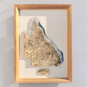 May include: A framed, three-dimensional map of Queensland, Australia, with a light wood frame. The map features layered, laser-cut wood showing the topography, with blue representing the ocean. A small plaque at the bottom reads "Queensland, Australia."