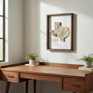 Könnte beinhalten: Eine gerahmte Holzkarte von Texas, die die Topographie des Staates mit einem detaillierten, geschichteten Design zeigt. Die Karte ist auf einem weißen Hintergrund in einem dunkelbraunen Rahmen montiert. Der Rahmen hängt an einer weißen Wand über einem Holzschreibtisch.