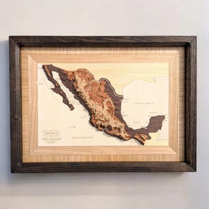 May include: A framed wooden map of Mexico with a 3D relief design. The map is made of different shades of wood to represent the different elevations of the country. The map is framed in a dark wood frame.