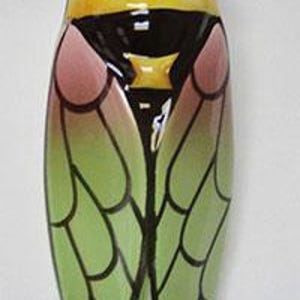 May include: A ceramic cicada wall hanging. The insect has a yellow and black head and thorax, with pink and green wings outlined in black. The cicada is approximately 6 inches long.