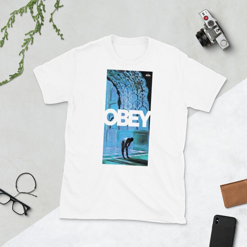 K&ouml;nnte beinhalten: Wei&szlig;es T-Shirt mit einem blauen und wei&szlig;en Grafikdruck einer Person, die in einem Durchgang steht, mit dem Wort "OBEY" in Wei&szlig; &uuml;ber der Person.