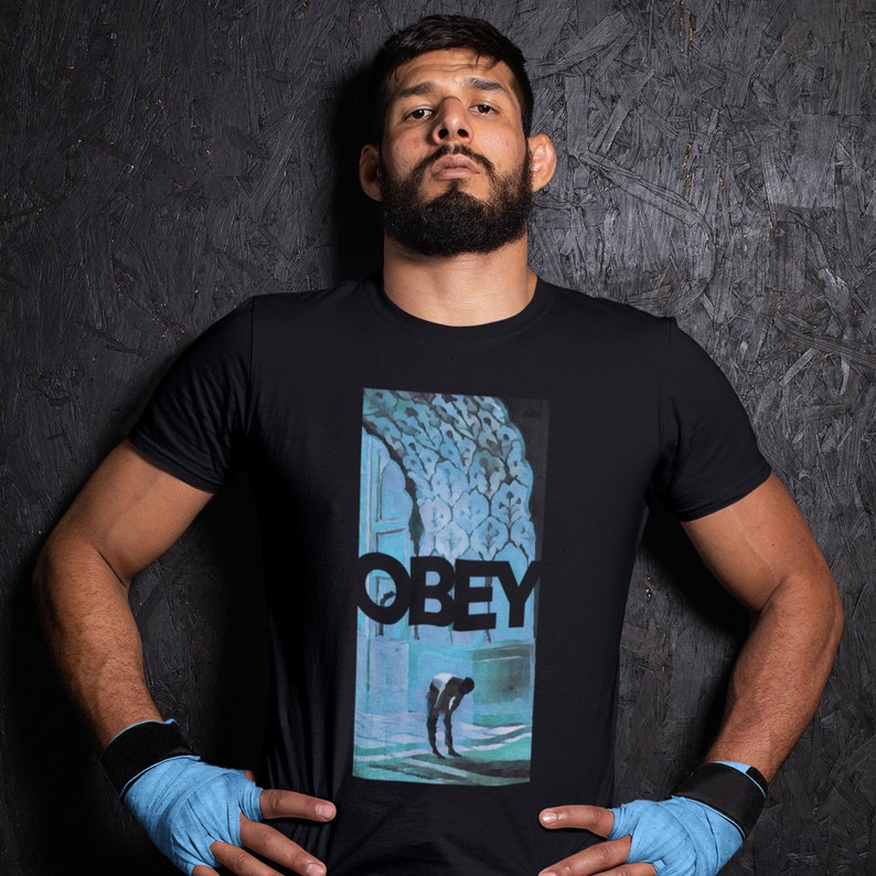 K&ouml;nnte beinhalten: Ein Mann tr&auml;gt ein schwarzes T-Shirt mit einem blauen und wei&szlig;en Grafikdruck. Die Grafik zeigt eine Person, die vor einer T&uuml;r steht, &uuml;ber der das Wort "OBEY" gedruckt ist. Der Mann tr&auml;gt an beiden Handgelenken blaue Armb&auml;nder.