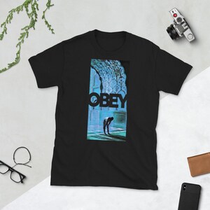 K&ouml;nnte beinhalten: Schwarzes T-Shirt mit einem grafischen Druck einer Person, die in einem blauen und gr&uuml;nen Raum steht. Der Text "OBEY" ist in schwarzen Buchstaben &uuml;ber der Person gedruckt.