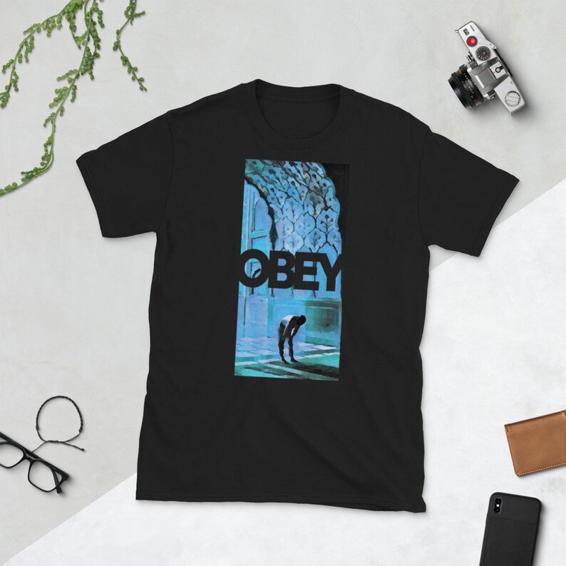 K&ouml;nnte beinhalten: Schwarzes T-Shirt mit einem grafischen Druck einer Person, die in einem blauen und gr&uuml;nen Raum steht. Der Text "OBEY" ist in schwarzen Buchstaben &uuml;ber der Person gedruckt.