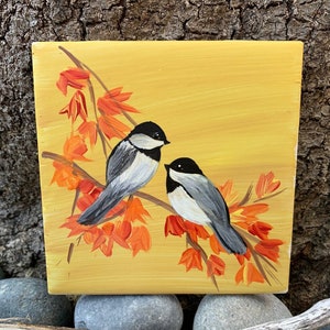 Puede incluir: Una obra de arte cuadrada pintada a mano que representa dos pequeños pájaros posados en una rama con hojas naranjas sobre un fondo amarillo. Los pájaros tienen marcas en blanco y negro. La obra de arte se exhibe sobre una superficie de madera natural.