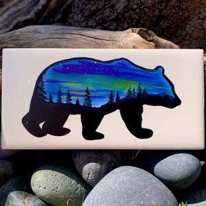 Könnte beinhalten: Eine handbemalte Keramikfliese mit der Silhouette eines schwarzen Bären, in dessen Innerem eine farbenfrohe Aurora Borealis-Szene dargestellt ist. Die Aurora ist in Blautönen, Lila, Grün und Gelb gemalt.