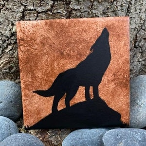 May include: A black silhouette of a wolf howling at the moon, painted on a small canvas with a rusty brown background.