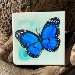 May include: A blue butterfly with black markings is painted on a white square tile. The tile is sitting on a piece of driftwood and surrounded by smooth, gray stones.