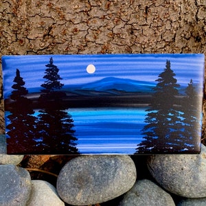 May include: A small painting of a nighttime scene with two dark green trees on either side of a blue lake. The moon is visible in the sky above the lake.