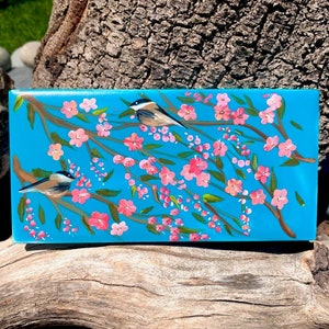 Può includere: Una piastrella dipinta a mano con uno sfondo blu che presenta due uccelli appollaiati su un ramo con fiori rosa. La piastrella è un pezzo decorativo per una casa.