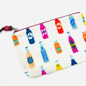 May include: A white zippered pouch with a red zipper pull and a brown strap. The pouch is decorated with a repeating pattern of colorful bottles with different labels, including "Sport", "Soda", "Fizzle", "Fresh", and "1UP". The pouch has the text "Floyd Clements" printed on the side.