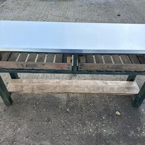 May include: A green metal table with a stainless steel top and two wooden crates underneath. The crates have the text "L.N. ADAMS" printed on them. The table has a wooden shelf underneath.