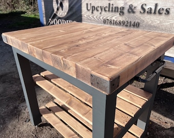 May include: A wooden kitchen island with a gray metal frame and two shelves. The top is made of reclaimed wood planks and has metal corner brackets. The island has a metal handle on the side.