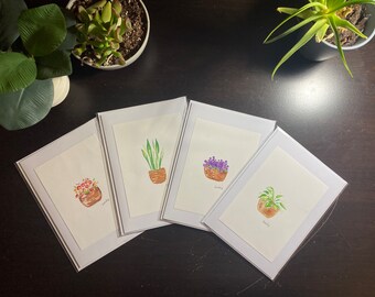 May include: Four watercolour paintings of potted plants on white card stock. The plants are a purple flower, a green plant with white flowers, a red flower, and a green plant with large leaves. Each painting is framed with a white border.