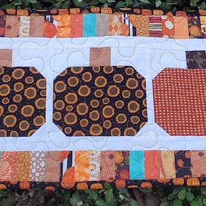May include: A rectangular quilted table runner featuring five pumpkin designs. Four pumpkins are brown with a circular pattern, and one is orange. The runner has a white center and a colorful border with orange, brown, and teal fabrics.