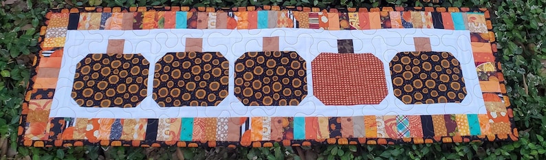 May include: A rectangular quilted table runner featuring five pumpkin designs. Four pumpkins are brown with a circular pattern, and one is orange. The runner has a white center and a colorful border with orange, brown, and teal fabrics.