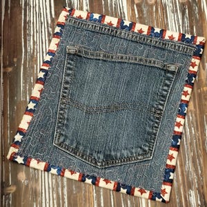 May include: A square denim placemat with a jean pocket and a patriotic red, white, and blue star border. The denim is a medium wash, and the border features alternating stars and stripes.